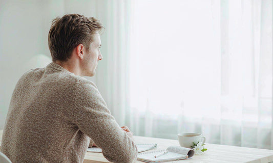 Man seated at a sunlit minimal desk with notebook and coffee seen from behind
