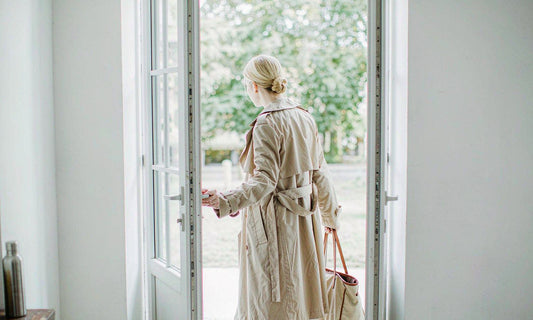 Woman seen from behind in a beige coat heading out the door with a tote bag and water bottle in morning light