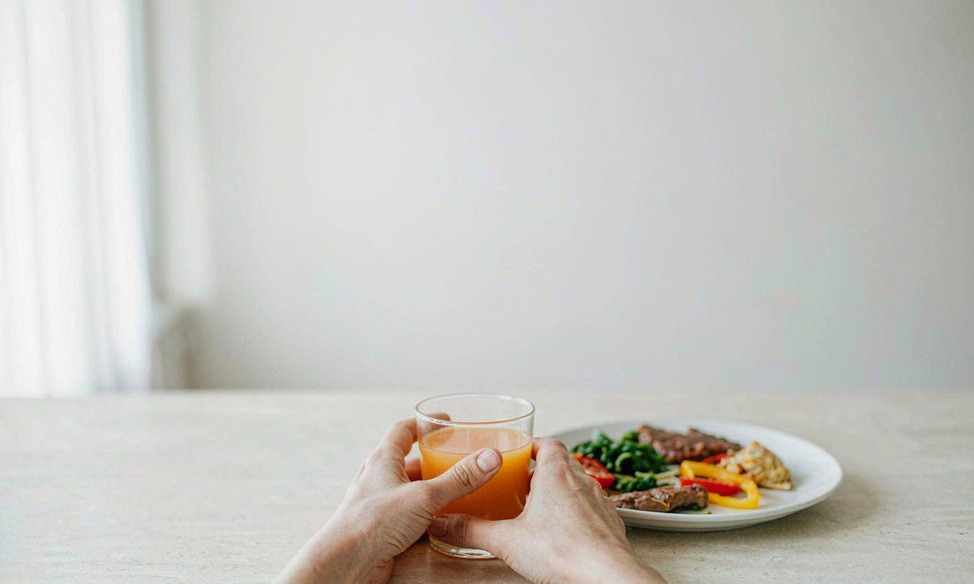 A pair of relaxed hands holding a small glass of orange juice beside a plain ceramic plate on a pale kitchen counter in soft window light
