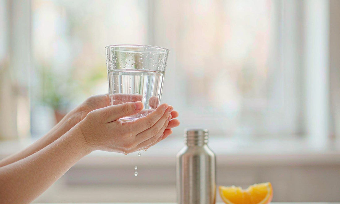 Caucasian hands with fair light skin holding a glass of water up to the light beside a steel bottle and sliced orange