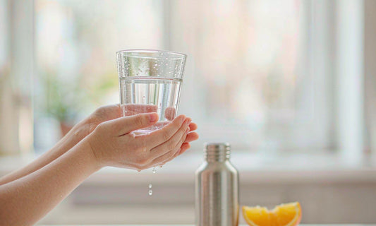 Caucasian hands with fair light skin holding a glass of water up to the light beside a steel bottle and sliced orange