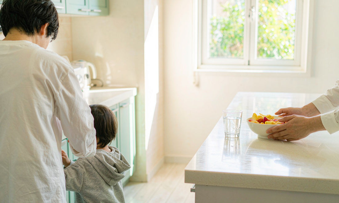 A softly out-of-focus parent and child seen from behind at a bright kitchen island with sliced fruit and a glass of water