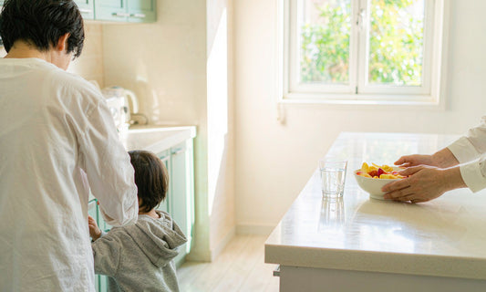 A softly out-of-focus parent and child seen from behind at a bright kitchen island with sliced fruit and a glass of water