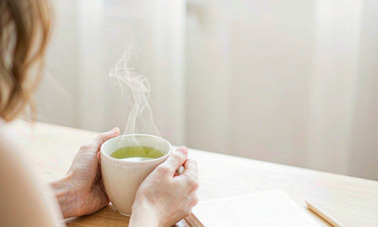 Hands cupping a steaming mug of green tea beside an open notebook on a wooden desk in soft daylight