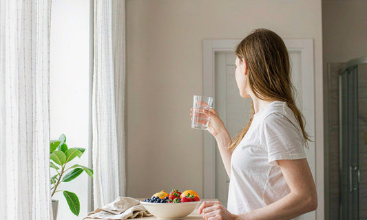 Woman seen from behind holding a glass of water beside a bowl of fresh fruit in a bright kitchen