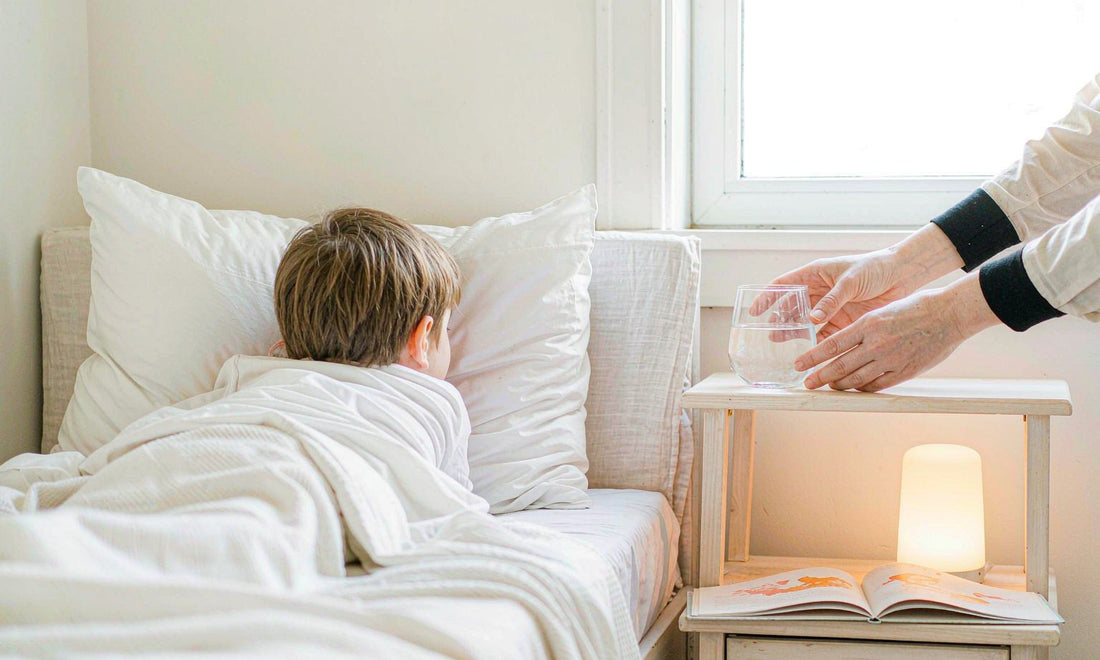 A softly out-of-focus child asleep in pale linen beside a bedside table with a warm nightlight, a storybook and a glass of water