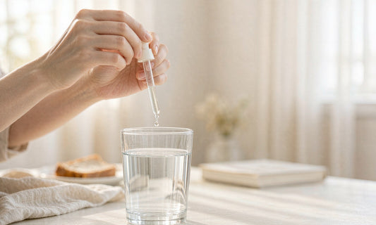 Hands holding a glass dropper above a clear glass of water on a sunlit breakfast table