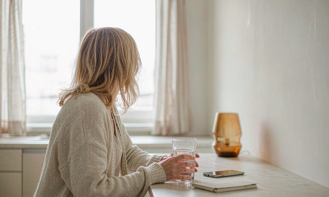 Woman seen from behind holding a glass of water in a bright kitchen during an early shift-work morning routine
