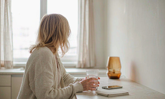 Woman seen from behind holding a glass of water in a bright kitchen during an early shift-work morning routine