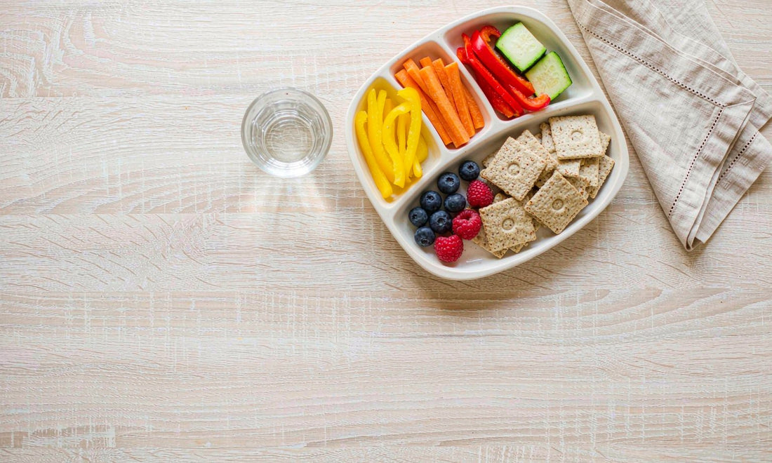 Top-down flat lay of a toddler meal with rainbow vegetables, berries and crackers on a pale oak table.