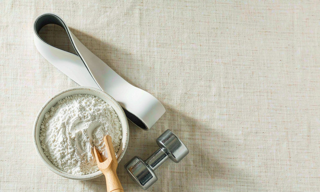 White creatine powder in a ceramic bowl with a wooden scoop beside a small dumbbell and resistance band
