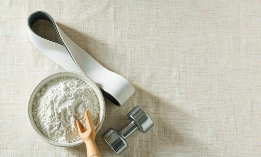 White creatine powder in a ceramic bowl with a wooden scoop beside a small dumbbell and resistance band