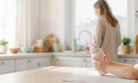 Light-skinned child's hands holding a pastel water bottle at a sunlit kitchen table with an adult nearby