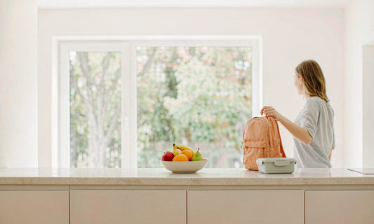 A woman seen from behind setting a backpack and lunch container on a sunlit counter