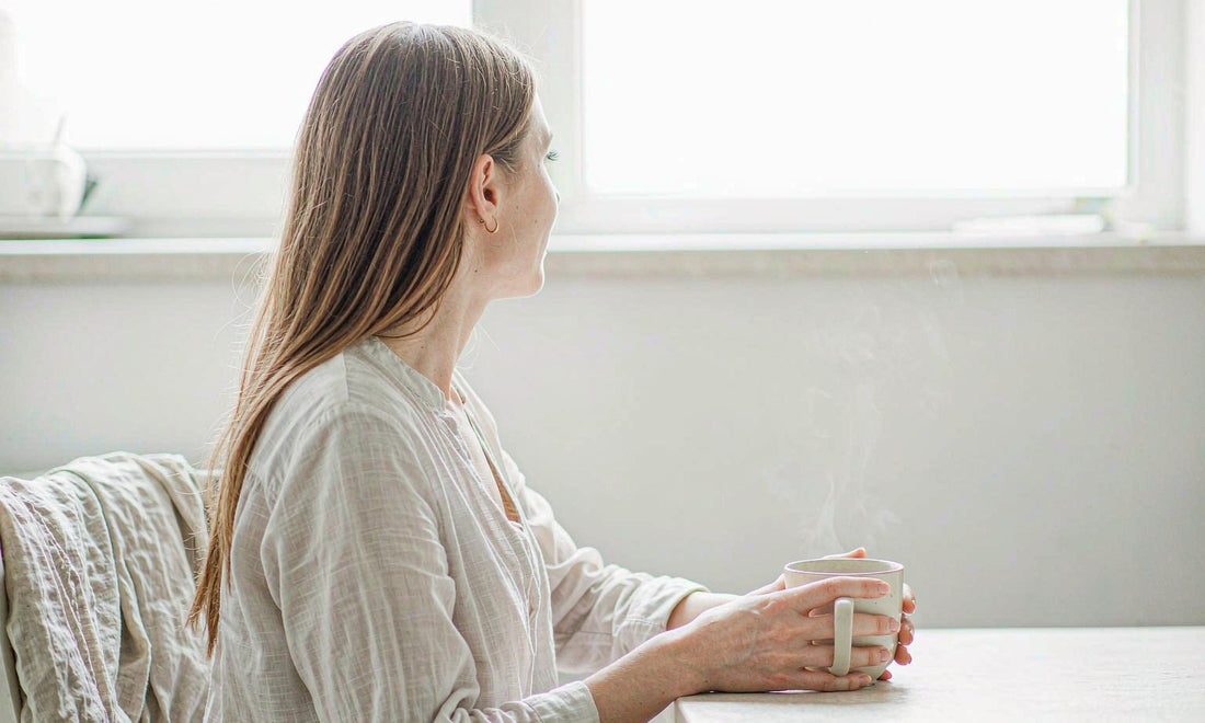 Woman cradling a warm mug of herbal tea at a pale evening kitchen table