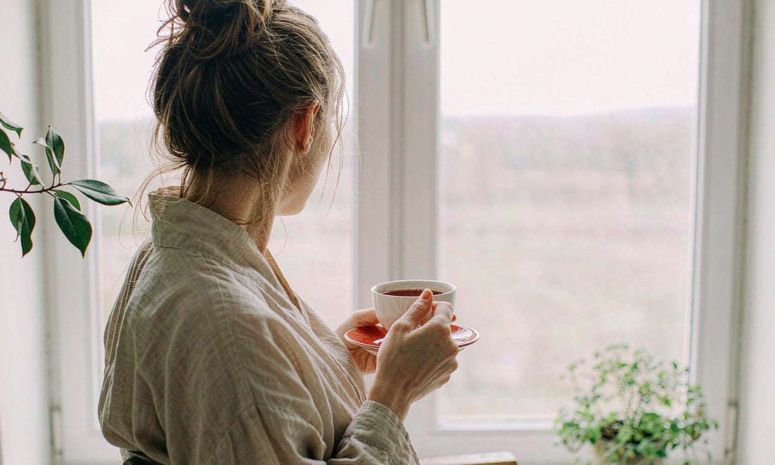 Woman seen from behind holding a warm mug by a bright morning window during a quiet daily routine