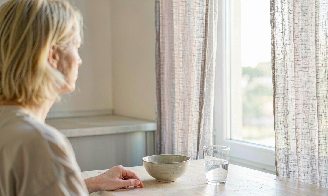 A woman with blonde hair sits at a pale kitchen table by a bright window beside an empty bowl and water