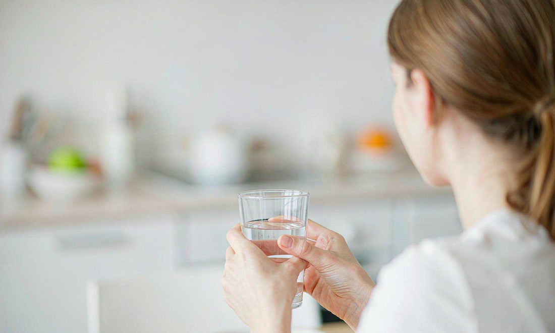 A woman seen from behind in a sunlit kitchen holding a glass of water in the early morning