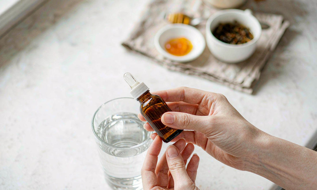 Fair skin hands holding an amber dropper bottle above a glass of water on a bright kitchen counter