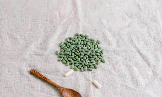 Flat lay of dried green seeds, white capsules and a wooden spoon on pale linen, supplement serving basics