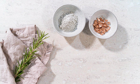 Top down flat lay of two dishes holding pale zinc powder and warm copper nuggets beside rosemary on stone
