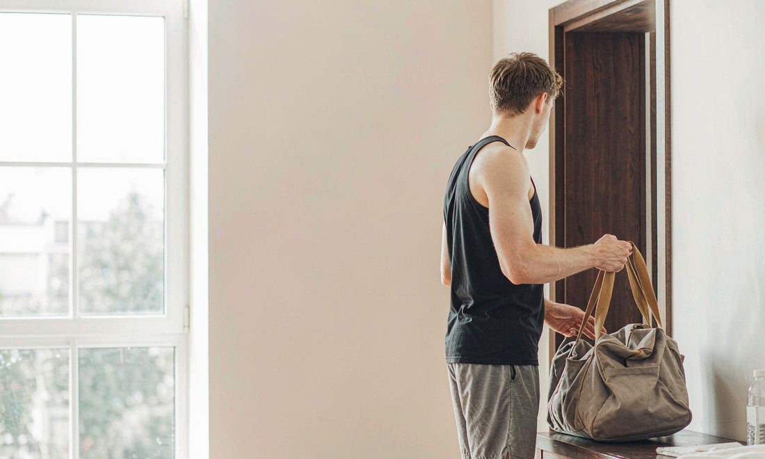 Man seen from behind lifting a canvas gym bag from a bench in a bright morning entryway