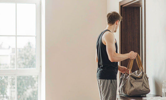 Man seen from behind lifting a canvas gym bag from a bench in a bright morning entryway