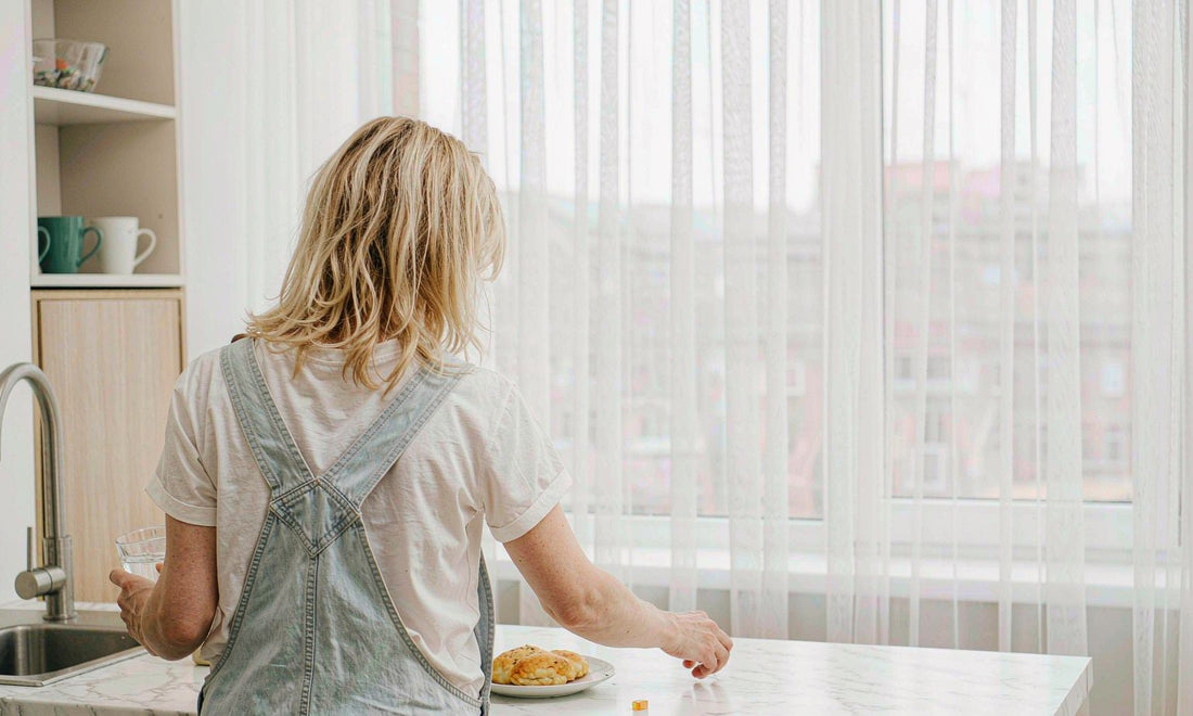 Woman with blonde hair seen from behind reaching for a gummy beside a glass of water in a bright morning kitchen