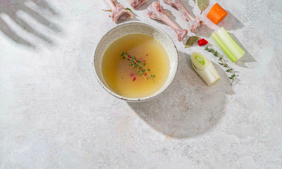 Overhead flat lay of a bowl of golden bone broth with fresh thyme, bones and vegetables on a pale counter