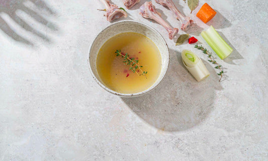 Overhead flat lay of a bowl of golden bone broth with fresh thyme, bones and vegetables on a pale counter
