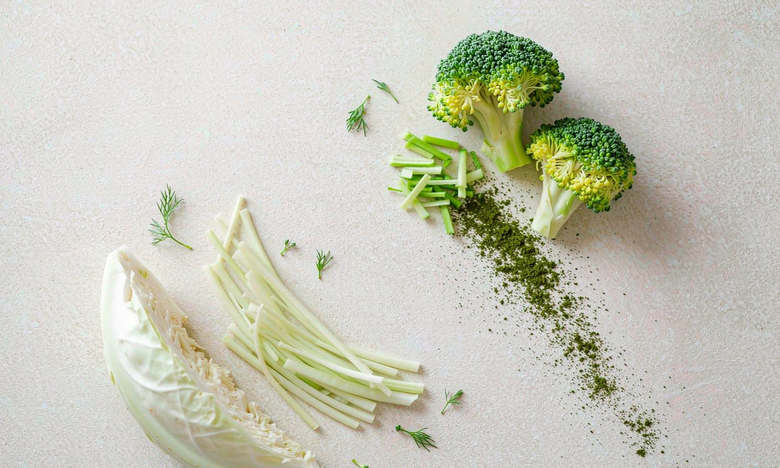 Overhead botanical arrangement of cruciferous greens, sliced stems, cabbage leaves and fine green powder on an off-white surface