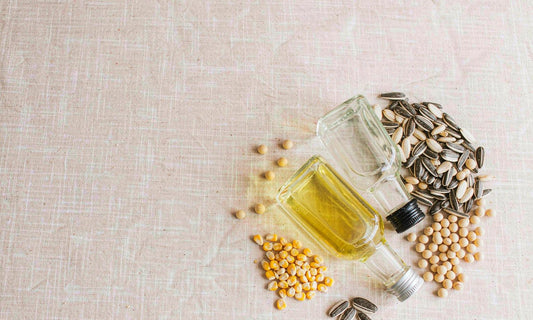 Overhead flat lay of glass oil bottles with sunflower seeds, soybeans and corn kernels on pale linen