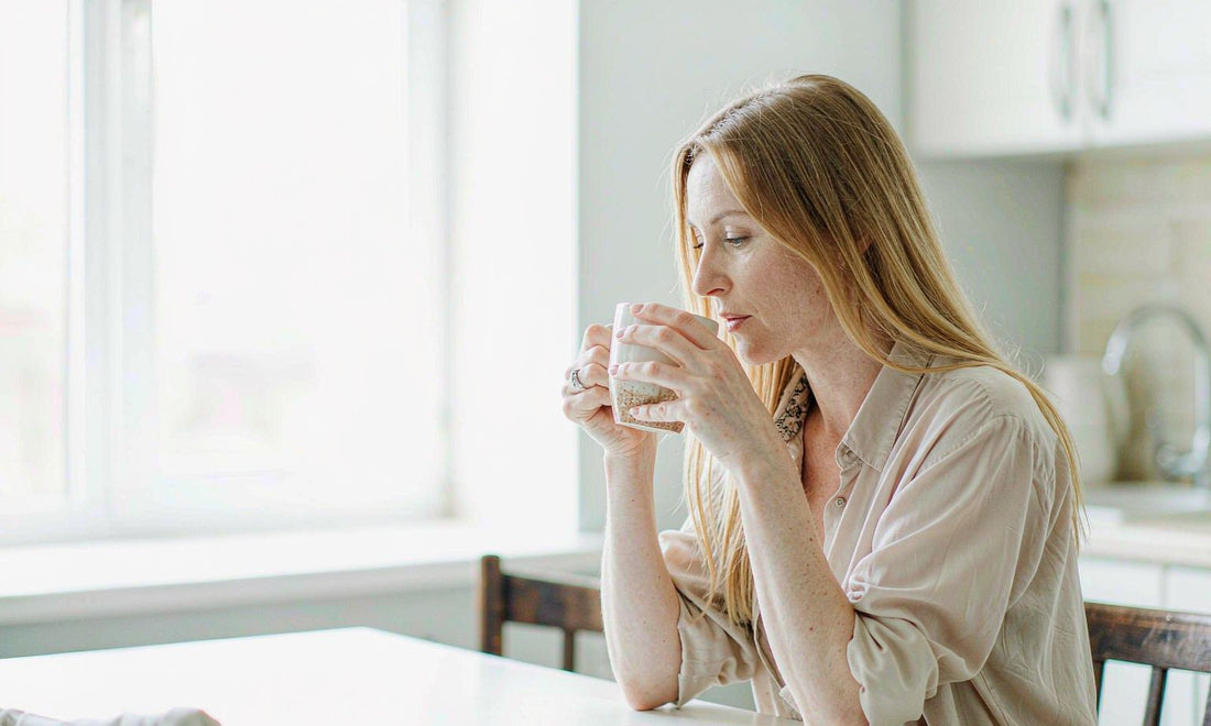 Woman seated at a bright kitchen table holding a mug, pausing with a warm drink in soft morning daylight