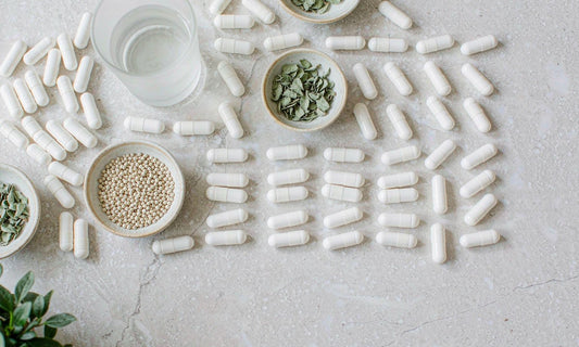 Overhead grid of plain white capsules, botanical leaves and water on pale stone