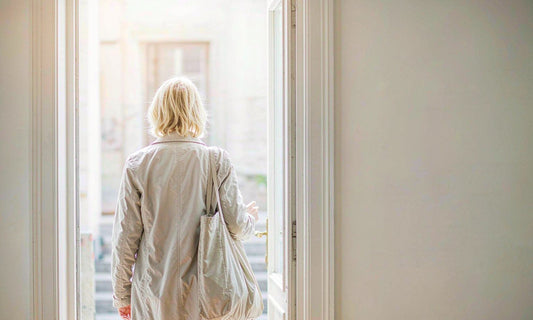 Person with light blonde hair seen from behind walking through a bright doorway with a tote bag