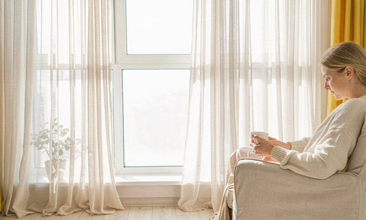 Western woman in cream sweater seen from side, holding a mug in sunlit window armchair