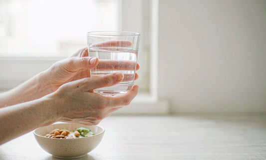Fair light skin hands lifting a glass of water from a sunlit kitchen counter beside a bowl of almonds