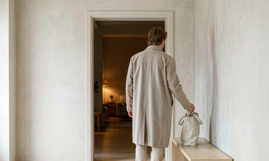 A man seen from behind sets down his tote bag on a wooden bench in a softly lit evening entryway