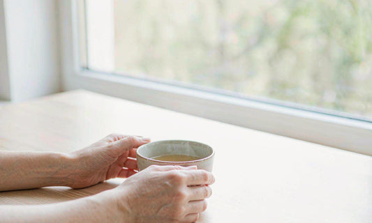 A pair of relaxed fair-skinned hands resting around a warm cup of tea by a window