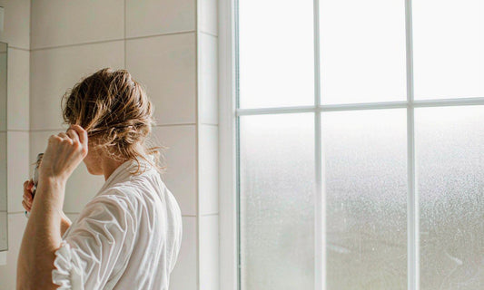 Woman with light brown hair seen from behind brushing her hair in a bright bathroom