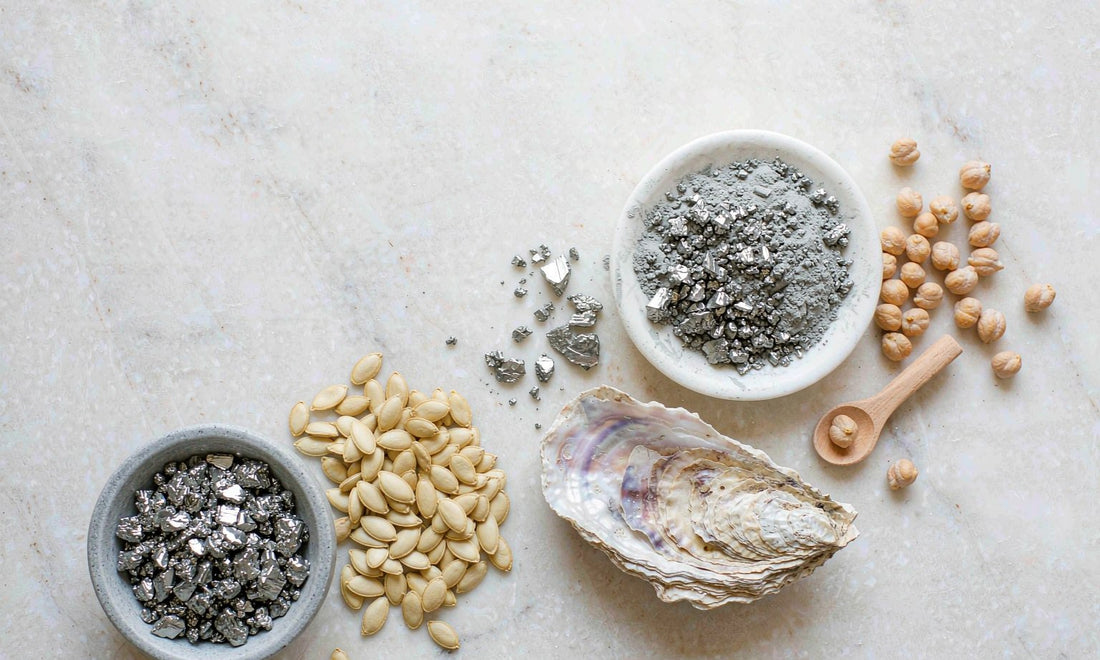 Flat lay of raw grey zinc mineral specimens, pumpkin seeds, chickpeas and oyster shell arranged on pale stone