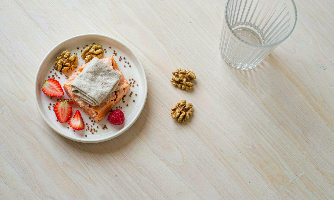 Overhead flat lay of a child plate with flaked salmon, walnut halves, berry slices and seeds on a light wooden table