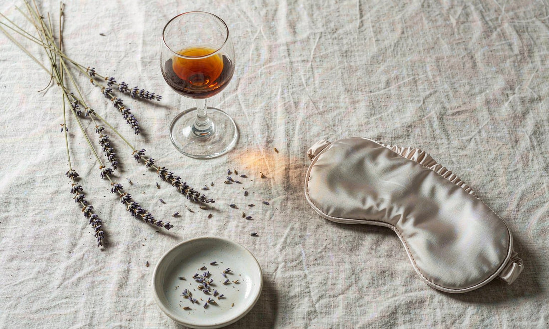 Overhead flat lay of a stemmed glass of amber liquid, dried lavender and a silk sleep mask on pale linen