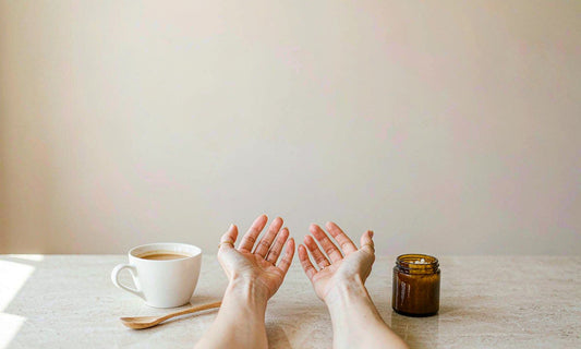 Relaxed hands beside a ceramic coffee cup and amber jar on a bright counter, morning light