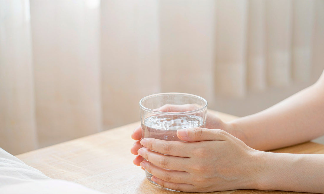 A pair of fair-skinned hands gently holding a glass of water on a pale wooden table at dawn.