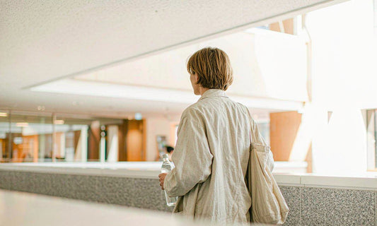 Person seen from behind walking through a bright lobby with a tote bag and water bottle