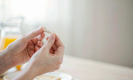 Hands holding a daily multivitamin capsule at a bright breakfast table in morning light